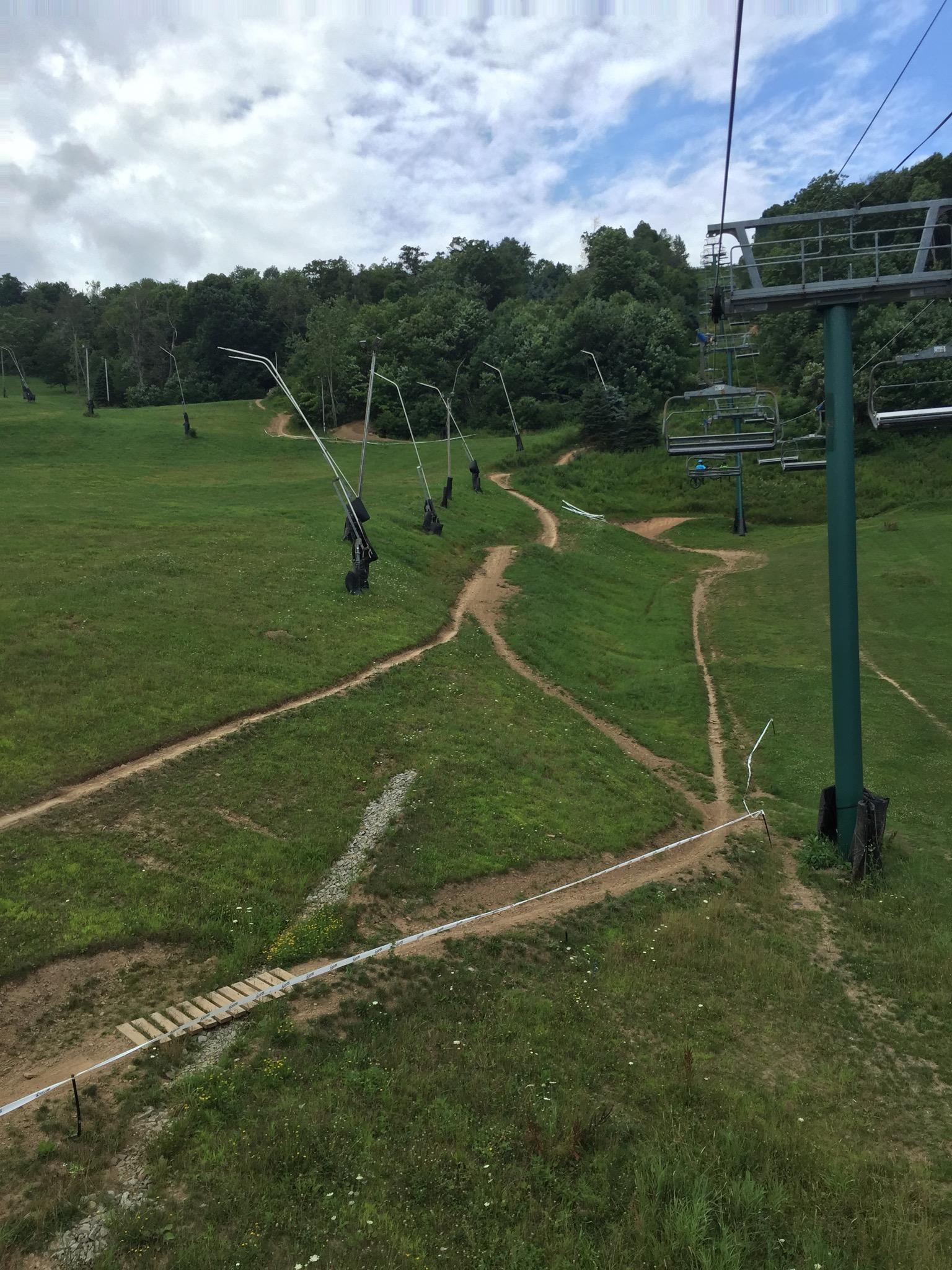 A view from a chairlift overlooking a grassy hillside with several ski lift towers in the background. The scene features winding dirt paths and patches of greenery, under a partly cloudy sky. Seven Springs mountain bike trail.
