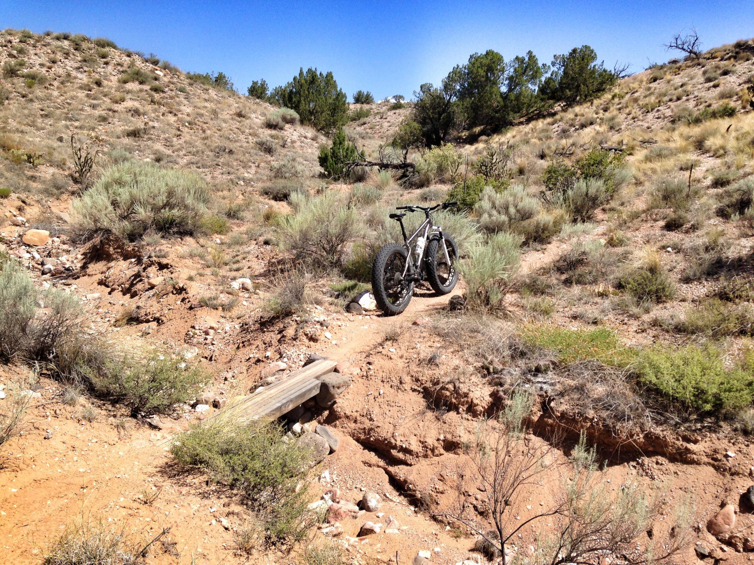 A fat-tire bike parked on a narrow dirt trail surrounded by dry, rugged terrain, with sparse vegetation and distant hills under a clear blue sky. Lower Reservoir trail mountain bike trail.