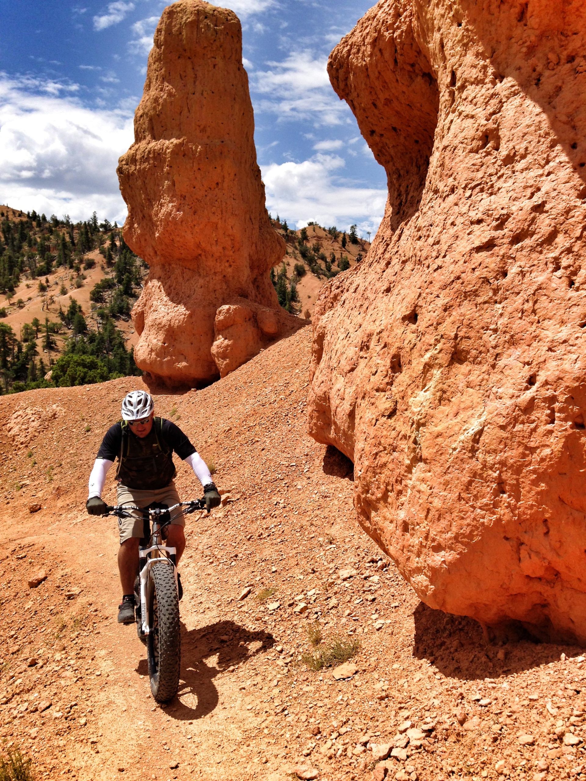A person riding a fat tire bike along a dirt path in a rugged landscape featuring tall, orange rock formations and blue skies with scattered clouds. Thunder Mountain mountain bike trail.