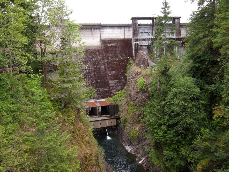 A large concrete dam stands at the top of a rocky gorge, surrounded by lush green trees. Water flows from the dam into the narrow canyon below. The dam features a textured surface, indicating age, with openings for water discharge. The scene captures the contrast between the man-made structure and the natural landscape. Wynoochee Lake mountain bike trail.