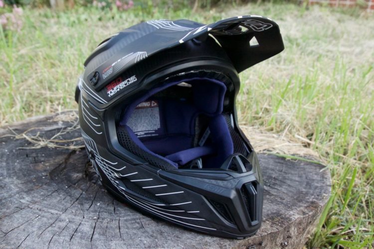 A black motocross helmet with a glossy finish and white graphic design sits on a wooden stump, surrounded by grass. The interior features purple padding for comfort, while the visor is slightly raised. The helmet showcases advanced protective technology, suitable for off-road biking.