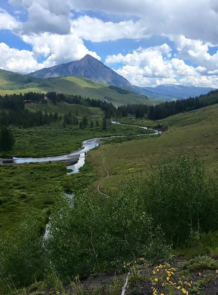 A scenic landscape featuring rolling green hills, a winding river, and a mountain peak under a partly cloudy sky. The foreground includes clusters of vegetation, and the background showcases expansive meadows and distant mountains. Lower Loop mountain bike trail.