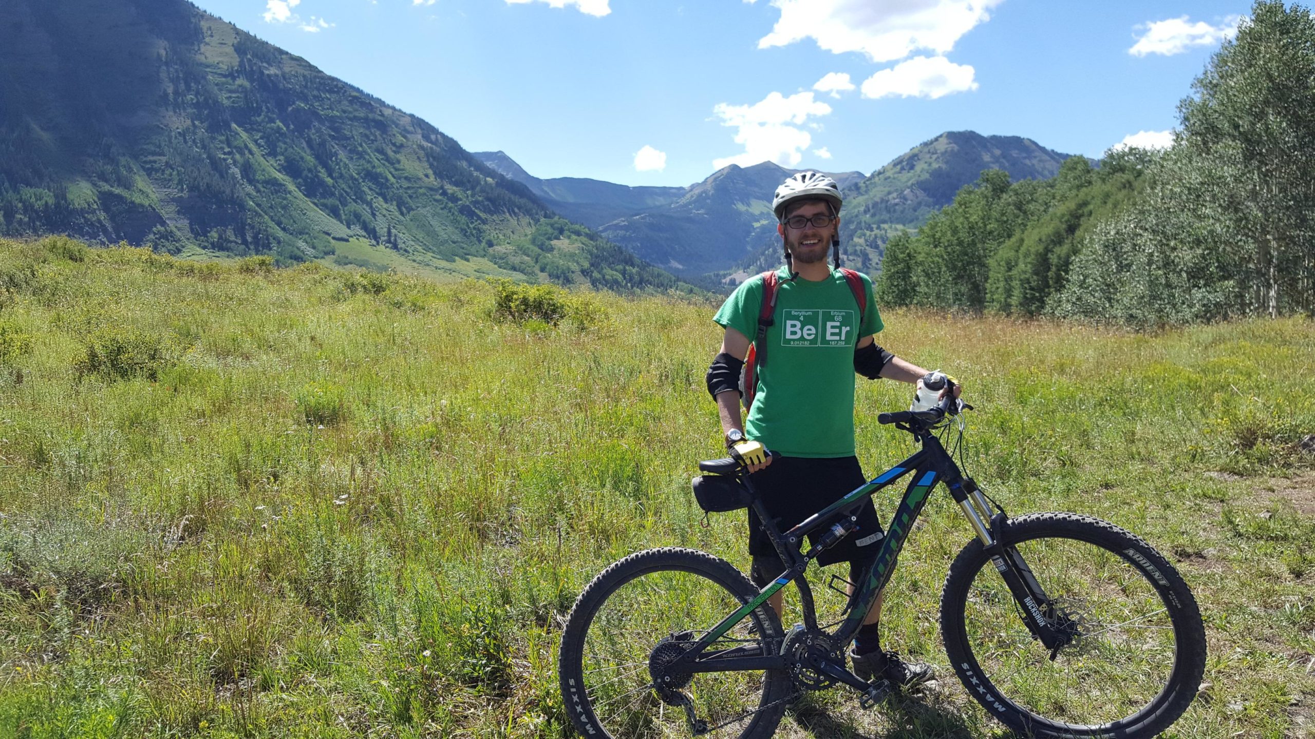 A person standing with a mountain bike in a grassy field surrounded by mountains, wearing a green t-shirt, black shorts, and bicycle gear, under a blue sky with some clouds. Lupine Trail mountain bike trail.