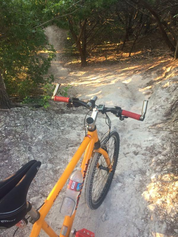 A close-up view of a mountain bike with orange frame and red handlebars, resting on a dusty dirt trail surrounded by trees. A bottle of water is attached to the bike frame, and the path ahead winds through the forest. Walnut Creek Trails mountain bike trail.