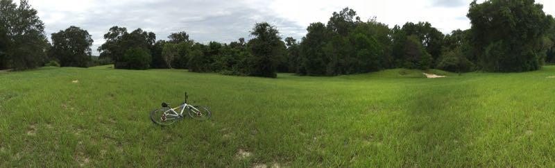 A panoramic view of a green field with tall grass, surrounded by trees. A bicycle lies on its side in the foreground, suggesting a moment of pause or abandonment in a natural, serene setting. The sky is overcast, adding to the tranquil atmosphere of the scene. Lake Apopka Restoration Area mountain bike trail.