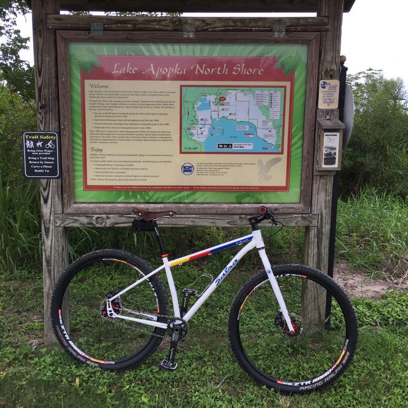 A mountain bike parked beside an informational sign for Lake Apopka North Shore, featuring a map of the area and details about the trails and safety tips for visitors. The background includes lush greenery and a hint of the lake. Lake Apopka Restoration Area mountain bike trail.