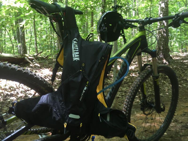 A mountain bike with a CamelBak hydration pack attached rests on a forested trail. The background features green foliage and sunlight filtering through the trees, creating a vibrant outdoor setting. Wild Turkey mountain bike trail.
