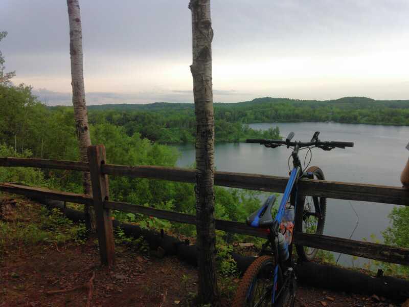 A scenic view overlooking a tranquil lake, surrounded by lush green trees and hills. In the foreground, a blue mountain bike leans against a wooden fence post, with a person partially visible on the right side of the image. The sky is overcast, suggesting a peaceful but moody atmosphere. Cuyuna Lakes mountain bike trail.