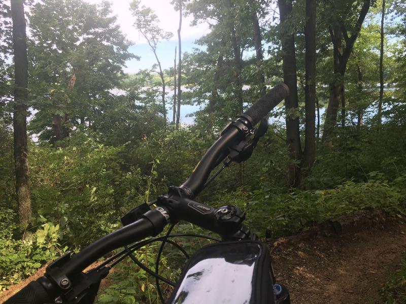 A mountain bike is positioned in the foreground, with its handlebars and control visible. In the background, a lush green forest surrounds a glimpse of a lake under a blue sky. The scene captures a serene outdoor environment, perfect for cycling or exploring nature. Fort Custer Recreation Area mountain bike trail.
