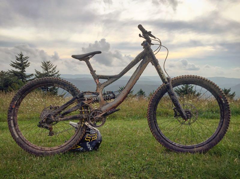 A mountain bike with a muddy frame and tires is resting on green grass, with a backdrop of rolling hills and a cloudy sky. A black helmet featuring the Fox logo is placed beside the bike. Snowshoe Bike Park mountain bike trail.