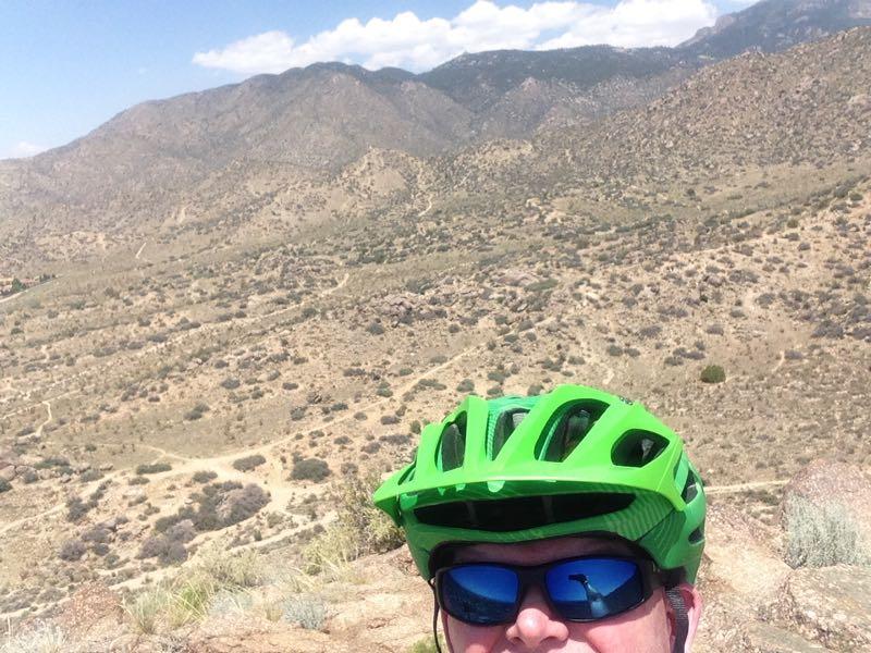 Selfie of a person wearing a bright green bike helmet and sunglasses, with a panoramic view of rugged mountains and a hilly landscape in the background. The sky is partly cloudy, indicating a sunny day for outdoor activities such as biking. South Foothills mountain bike trail.