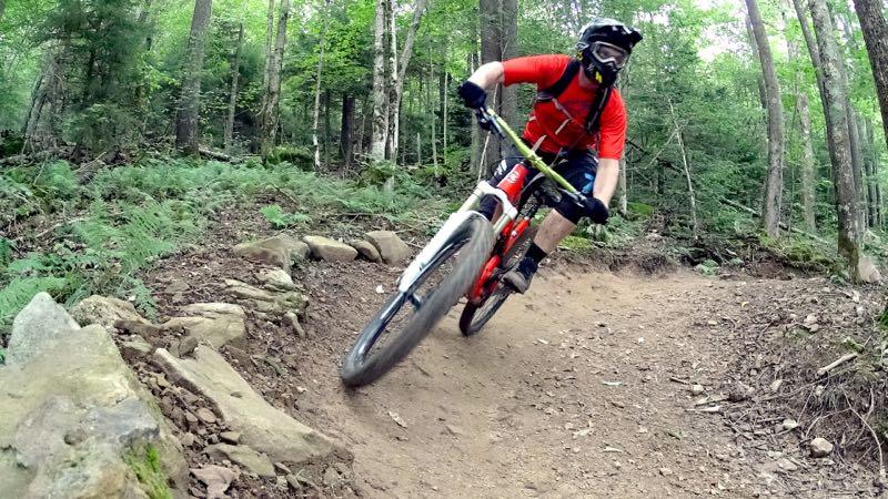 A mountain biker in a red shirt and helmet leans into a curve on a dirt trail surrounded by lush green trees and ferns. The bike features a white front wheel and is actively navigating a rocky path. Snowshoe Bike Park mountain bike trail.