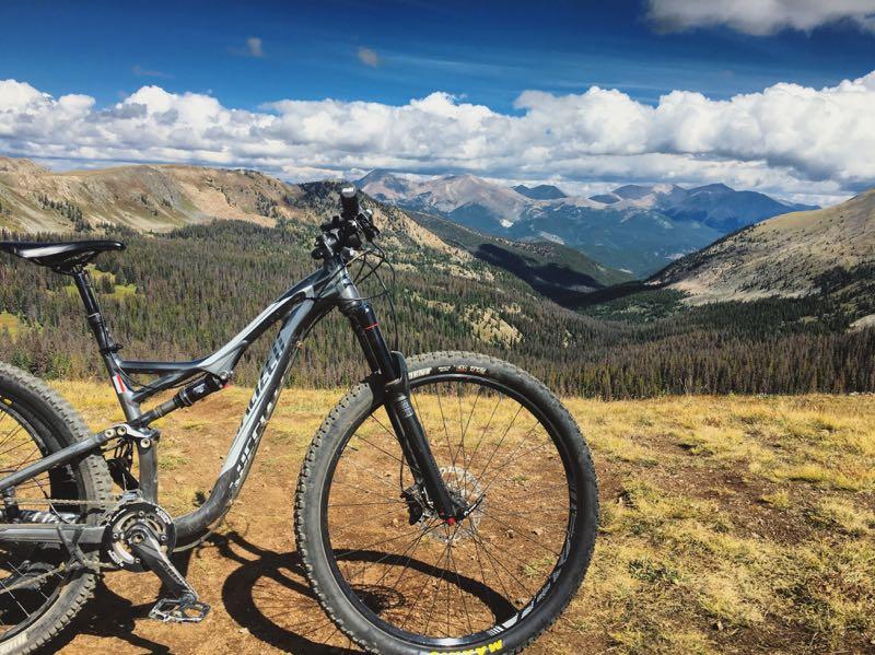 A mountain bike is positioned on a grassy ridge, overlooking a vast landscape of rolling hills, valleys, and distant mountains under a partly cloudy sky. The scene captures the beauty of nature, highlighting the adventurous spirit of cycling in a mountainous terrain. Monarch Crest Trail mountain bike trail.