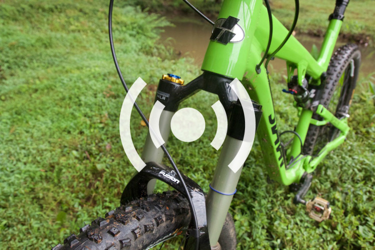 Close-up of a green mountain bike's front suspension fork and tire, set against a background of lush green grass. The bike features visible branding on the fork and a rugged tire tread pattern.