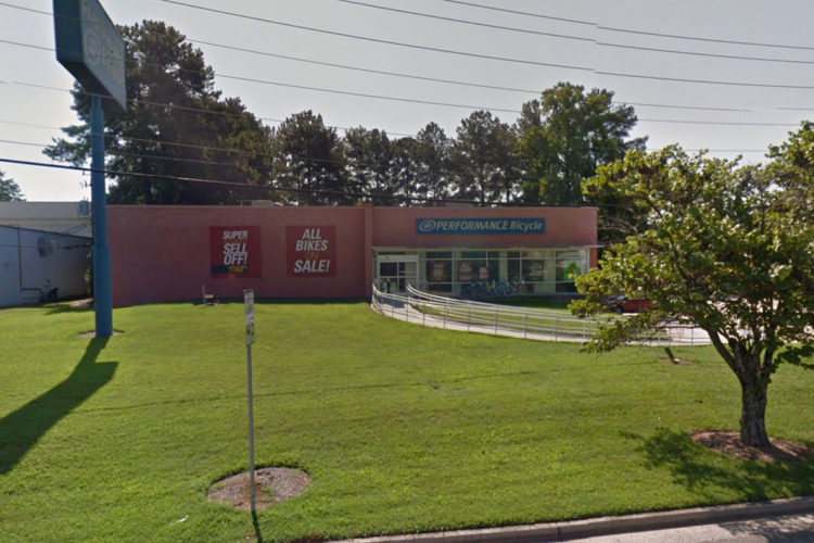 Exterior view of a Performance Bicycle store featuring large signage advertising "Super Sale!" and "All Bikes on Sale!" The building is set in a green grassy area with trees and power lines in the background.