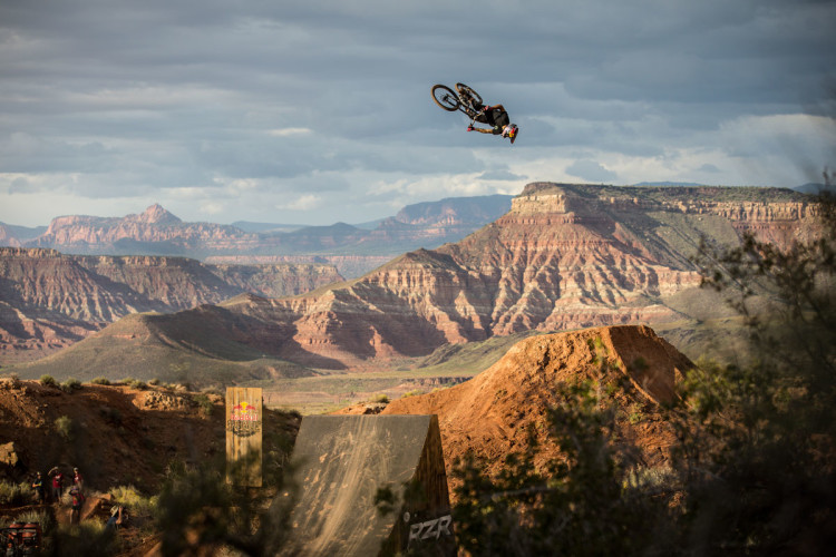 A mountain biker performing a backflip in mid-air above a dirt jump, set against a dramatic landscape of layered red rock formations and mountains under a cloudy sky. A crowd can be seen in the lower left, observing the stunt. The scene captures the thrill of extreme sports in a picturesque outdoor setting.