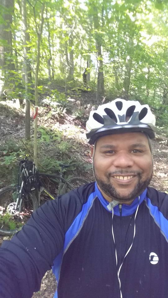 A person smiling while wearing a bicycle helmet and a blue athletic shirt, standing in a wooded area. A mountain bike is parked nearby, surrounded by greenery and trees in the background. Rosaryville State Park mountain bike trail.