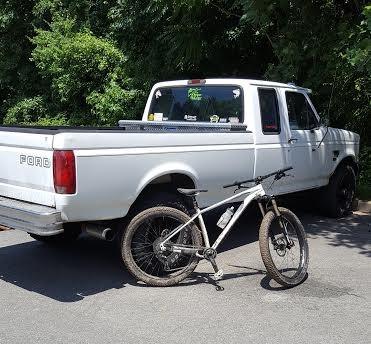 Specialized Fuse Comp 6Fattie: A white Ford pickup truck parked next to a mountain bike on a sunny day, surrounded by green foliage.