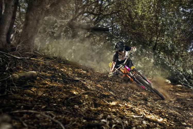 A mountain biker skillfully navigating a dirt trail, leaning into a turn with dust and debris flying around. The rider is wearing a helmet and protective gear, surrounded by trees and rocks in a natural outdoor setting.