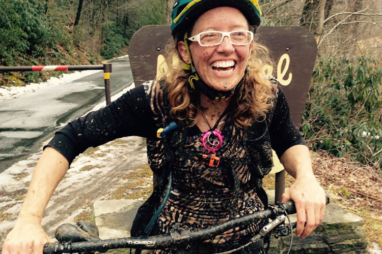 A smiling person with curly hair, wearing a colorful helmet and glasses, stands beside their bicycle, which is muddy from a recent ride. They are covered in dirt, indicating a fun and adventurous outdoor activity, with a wooded area and a road visible in the background.