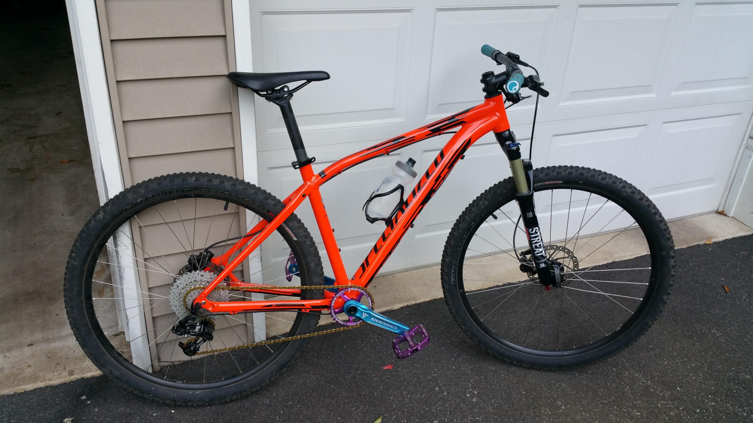 Specialized Pitch: A bright orange mountain bike with a sleek design, featuring wide tires and a purple pedal. It is positioned next to a garage door, showcasing its lightweight frame and components, including a water bottle holder and gears. The setting appears to be a driveway with a concrete floor.