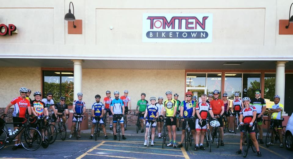A group of cyclists gathered in front of Tomten Biketown, smiling and posing for the photo. They are wearing various cycling jerseys and helmets, standing with their bicycles. The store