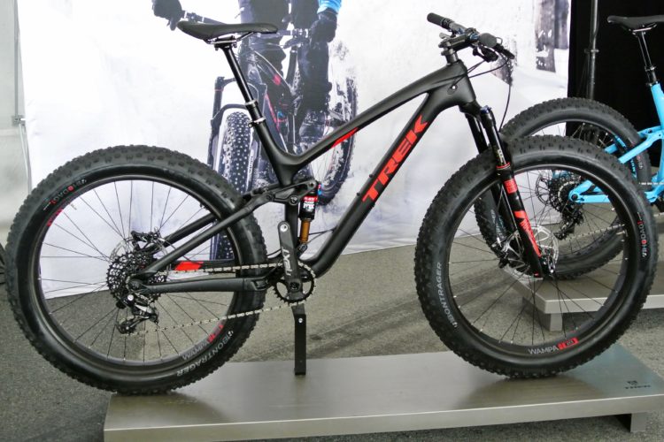 A sleek Trek fat bike displayed on a stand, featuring a black frame with red accents and wide, durable tires. The background includes a blurred image of cyclists mountain biking, emphasizing an outdoor adventure theme.