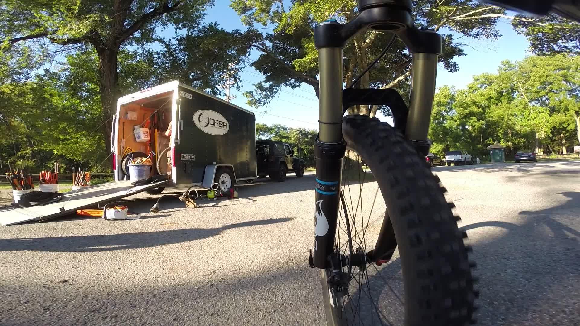 Close-up view of a mountain bike's front wheel, with a trailer in the background. The trailer has its doors open, revealing various tools and equipment, while bright sunlight filters through surrounding trees. The scene is set in a gravel parking area with a few parked vehicles visible. Allaire State Park mountain bike trail.