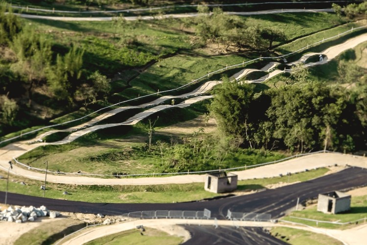 Aerial view of a winding road surrounded by lush green landscape, with pathways curving through trees and open fields. The scene includes a few small structures along the road and various segments of paved and unpaved surfaces.