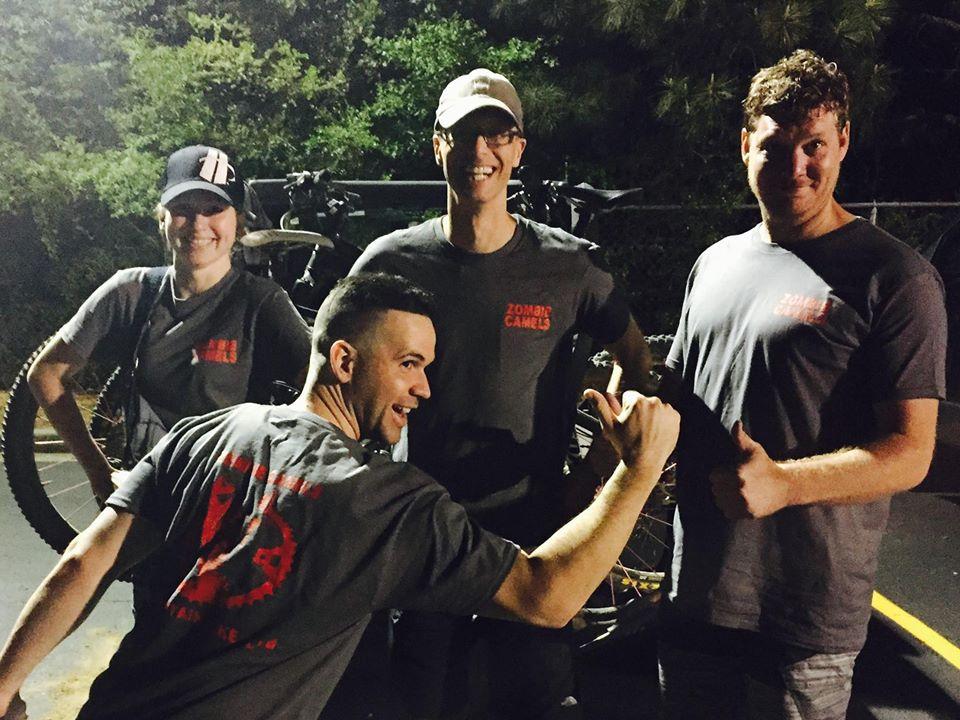 Four individuals pose together in a parking area at night, wearing matching gray shirts with the text "ZOMBIE CAMELS." They are smiling and displaying a variety of playful poses, with one person giving a thumbs up, while another strikes a fun, exaggerated pose. In the background, bicycles are visible, and lush greenery can be seen surrounding the area.