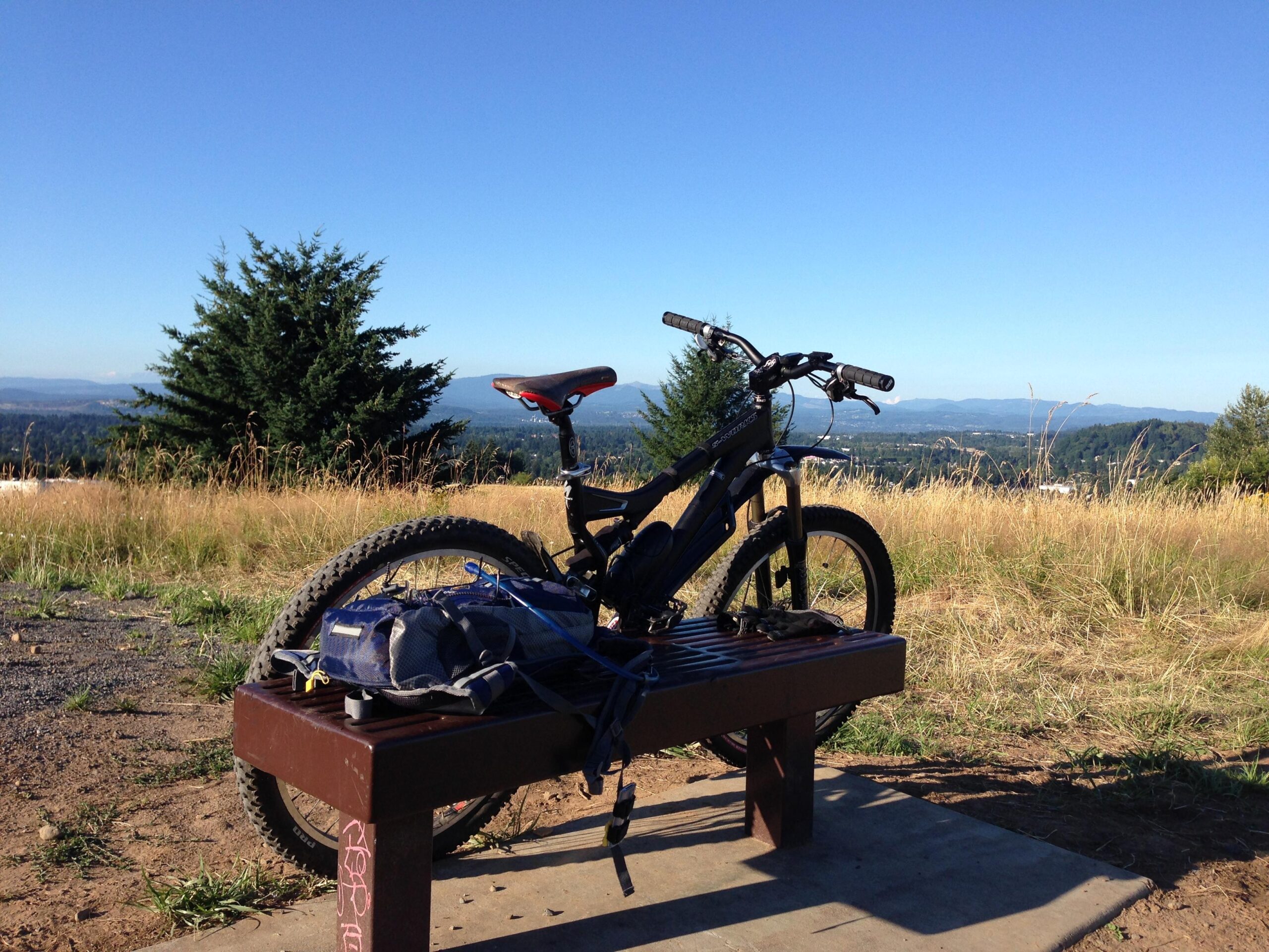 Specialized S-Works Stumpjumper FSR: A mountain bike rests on a wooden bench in a grassy area, with a view of rolling hills and mountains in the distance under a clear blue sky. A backpack is also placed on the bench beside the bike.