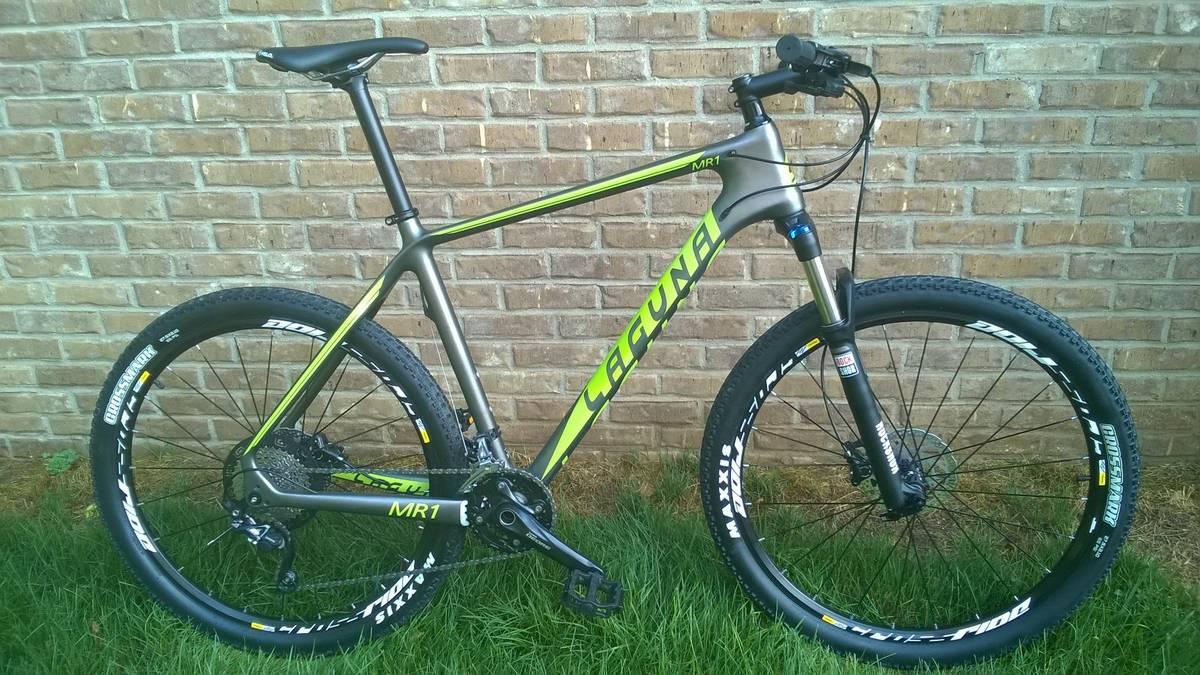 Laguna MR1: A modern mountain bike with a matte black and bright green frame, featuring the model name "MR1." The bike has thick, knobby tires designed for off-road terrain and is positioned on grass in front of a textured brick wall.