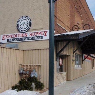 Exterior view of "Expedition Supply," a sporting goods store featuring a sign that highlights their offerings of ski and cycling gear. The building has a wooden facade and a snowy sidewalk, with a bicycle mounted on the roof.