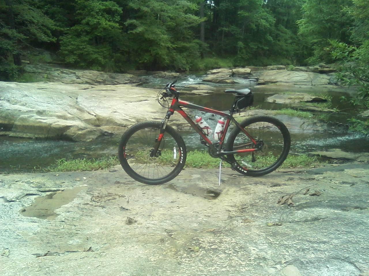 Mountain bike parked on rocky terrain near a flowing river, surrounded by lush green trees. Two water bottles are mounted on the bike frame. Cochran Mill Park mountain bike trail.