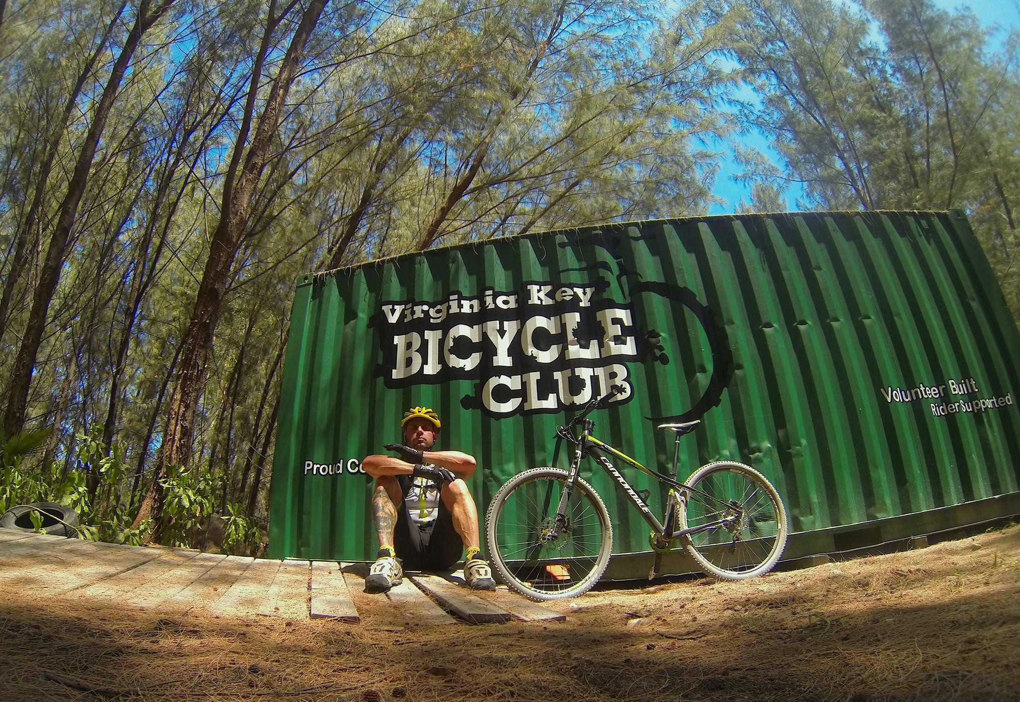 A cyclist sitting on a wooden platform next to a mountain bike, with a green shipping container featuring the text "Virginia Key BICYCLE CLUB" in the background. Tall trees are visible on either side, indicating a natural outdoor setting. The cyclist has a yellow helmet and a relaxed demeanor. Virginia Key North Point mountain bike trail.