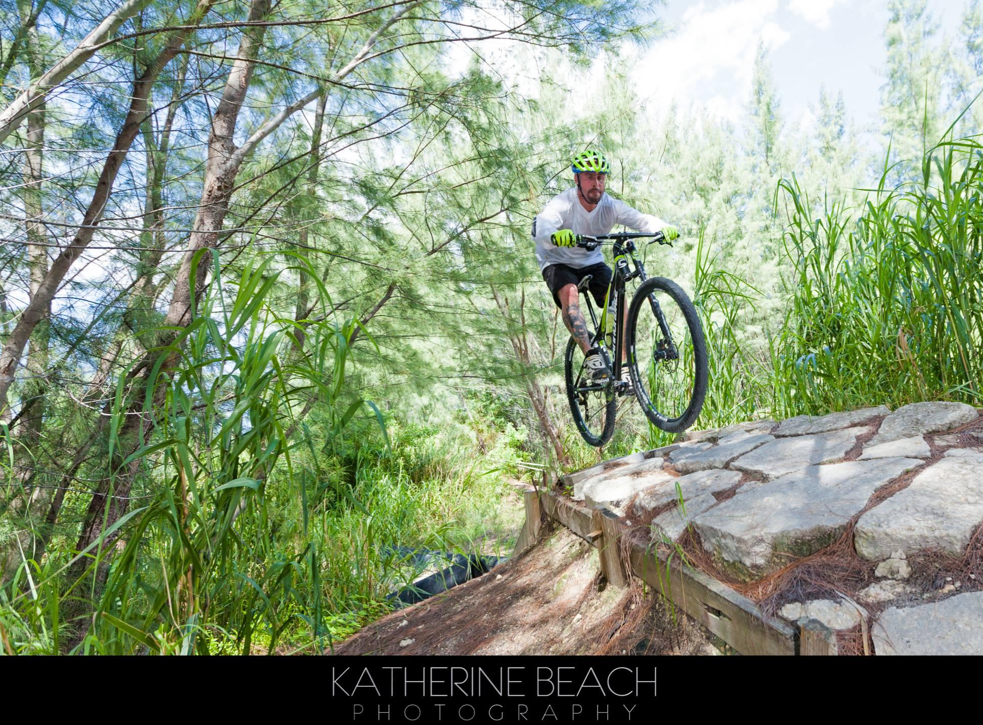 A mountain biker in a bright green helmet and gloves is captured mid-air as he jumps off a stone pathway surrounded by lush greenery and trees. The scene showcases the excitement and skill of mountain biking in a natural environment. Virginia Key North Point mountain bike trail.