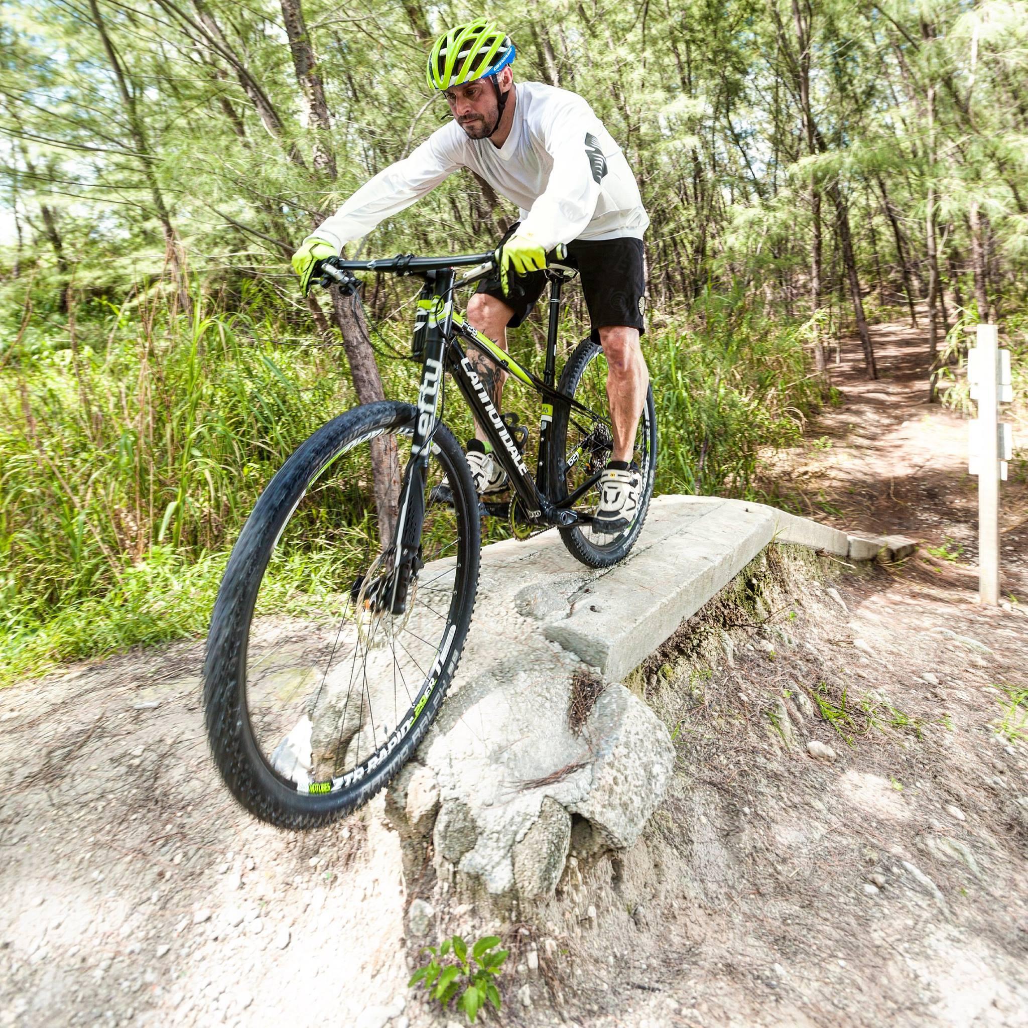 A cyclist riding a mountain bike over a rocky path in a forest. The rider is wearing a white long-sleeve shirt, black shorts, and a bright green helmet and gloves. The bike is in motion, capturing the dynamic action of mountain biking on a rugged trail surrounded by trees and greenery. Virginia Key North Point mountain bike trail.