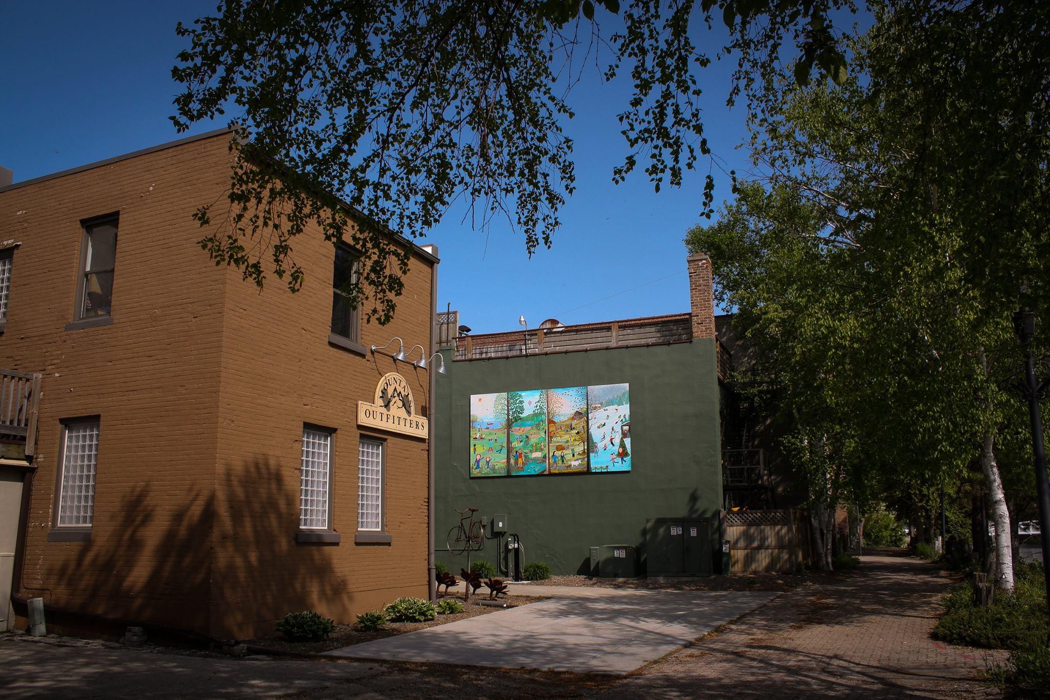 A street view of two buildings: a brown brick structure on the left with a sign that reads "Mountain Outfitters," and a green building on the right featuring a large, colorful mural. The street is lined with trees, and a pathway runs alongside the buildings. Clear blue skies overhead.
