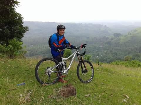 Giant Giant Trance 27.5 2: A person in a blue and orange cycling jersey and a helmet stands next to a mountain bike on a grassy hilltop, with a lush green landscape and distant hills in the background.