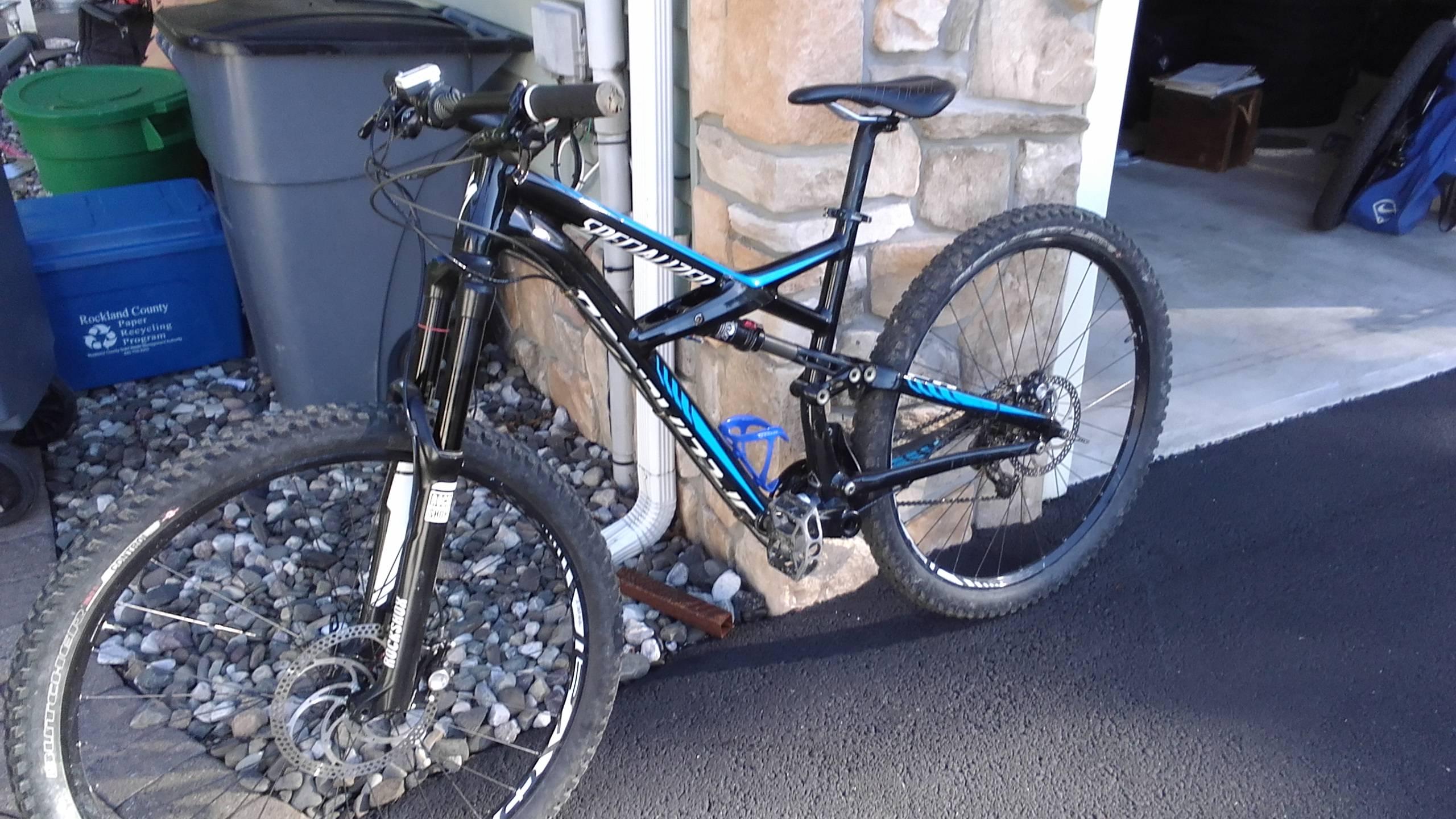 Specialized Enduro Comp 29: A black and blue mountain bike parked on a driveway, with gravel and recycling bins in the background. The bike features thick tires and a visible disc brake. A stone wall and a garage door are partially visible.
