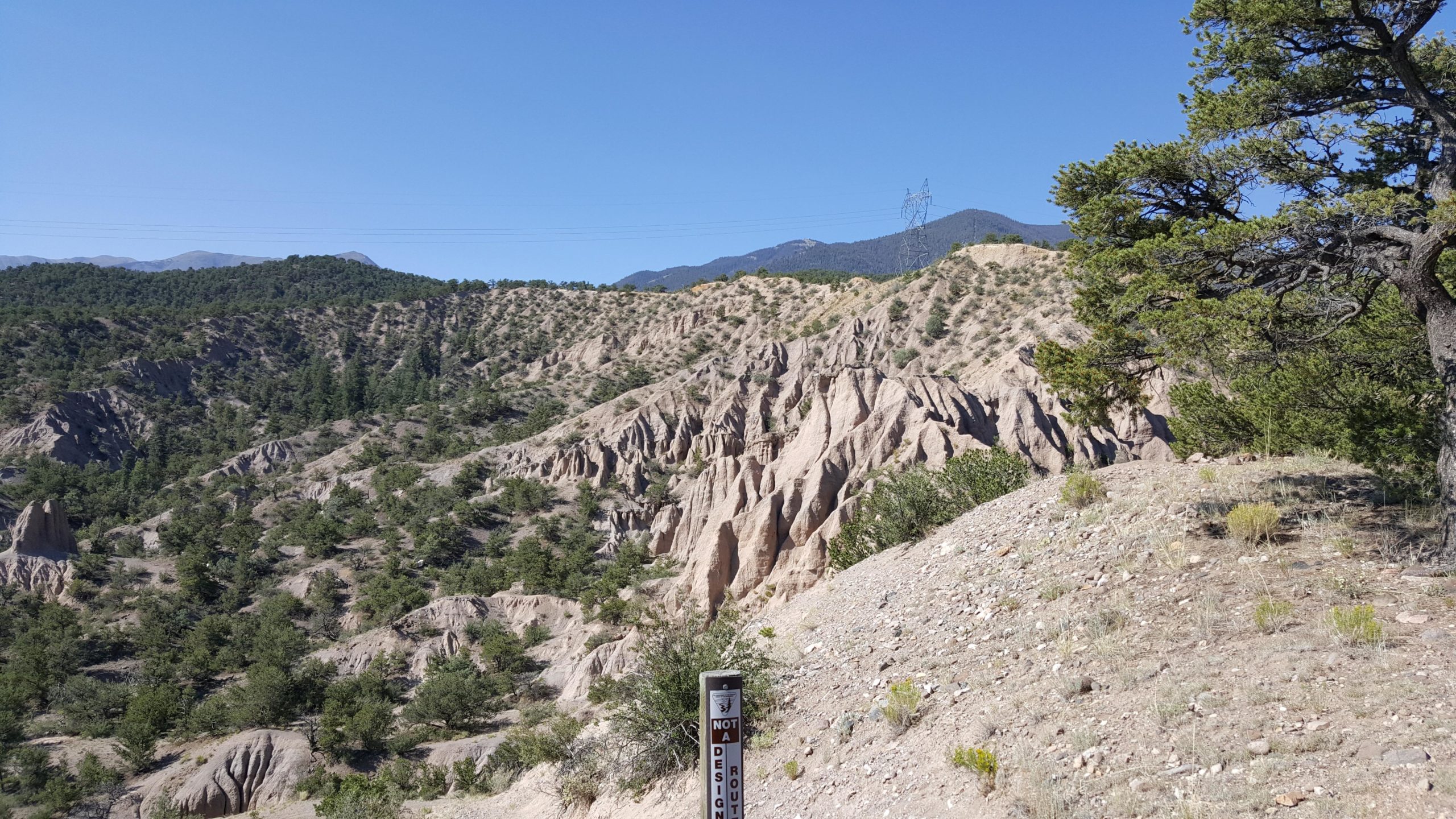 A scenic landscape featuring rugged terrain with steep, sloping hills covered in sparse vegetation. In the foreground, a sign marked "NOT A DESIGNATED ROUTE" stands near the edge of the hillside. The background showcases a mountain range under a clear blue sky, with scattered trees in the area. Little Rainbow mountain bike trail.