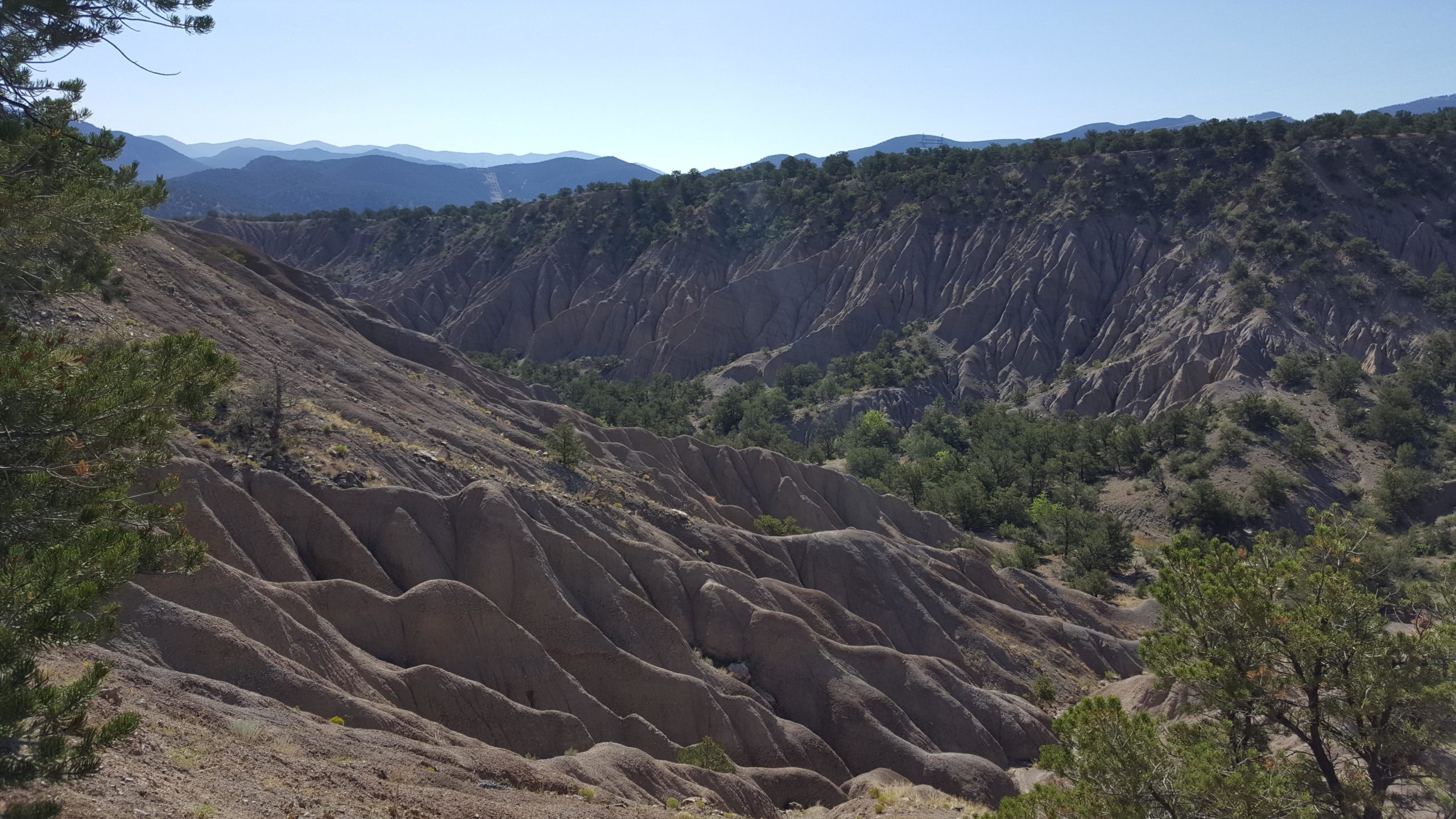 A panoramic view of rugged, sloping terrain characterized by soft, rolling hills and steep ridges. The landscape features light-colored, eroded rock formations and scattered patches of green vegetation, with distant mountains visible in the background under a clear blue sky. Little Rainbow mountain bike trail.