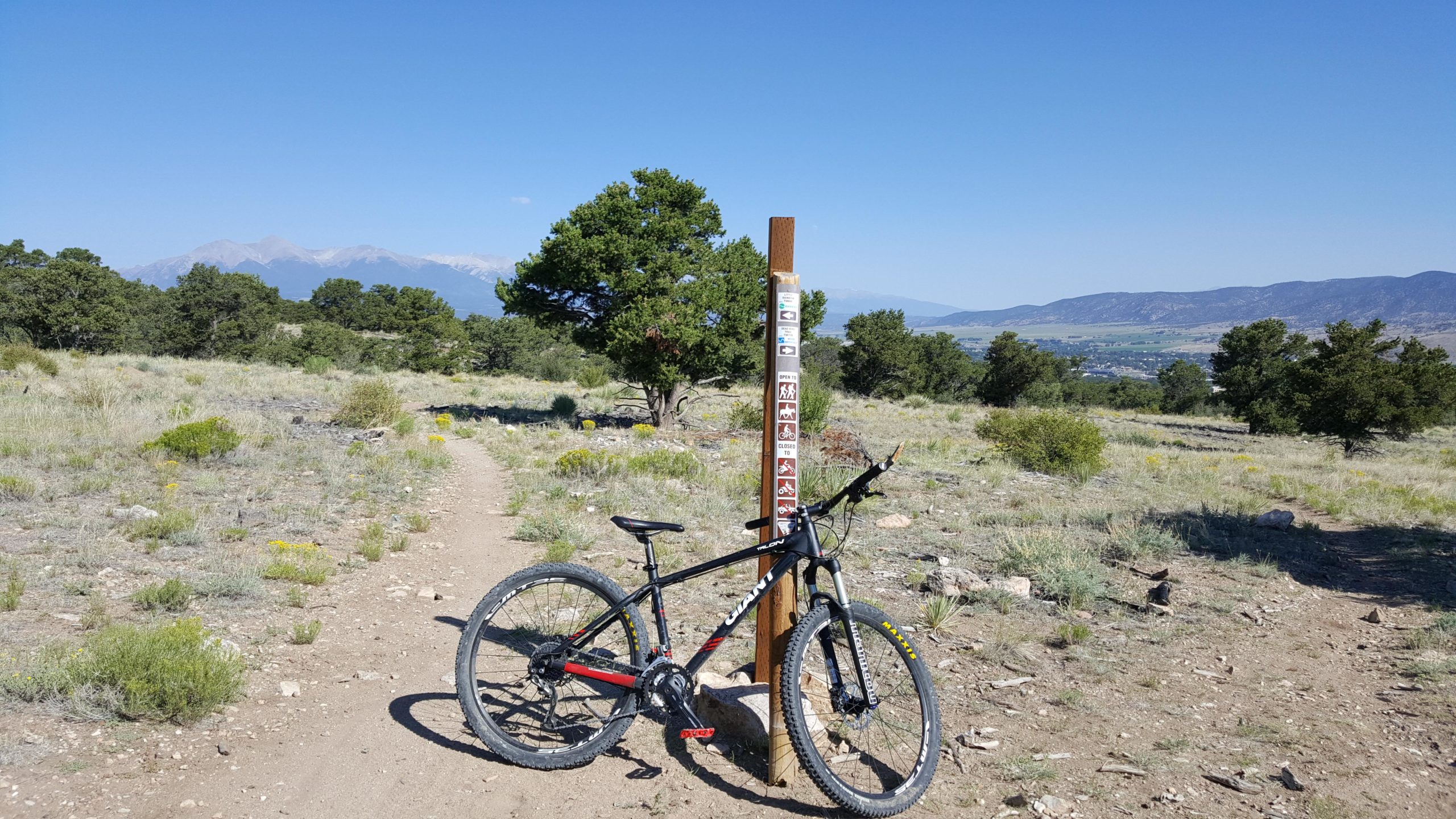 A mountain bike leaning against a trail marker on a dirt path surrounded by grassy terrain and sparse trees, with mountains visible in the background under a clear blue sky. Little Rainbow mountain bike trail.