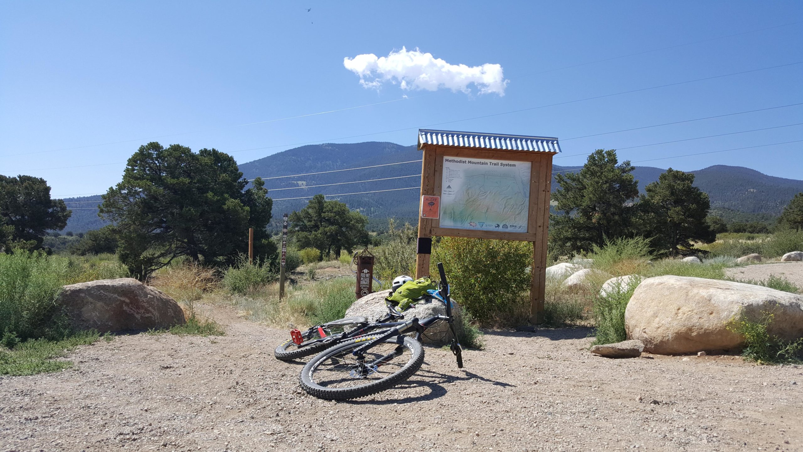 A mountain bike rests on the ground in front of a trailhead sign for the Methodist Mountain Trail System, surrounded by rocky terrain and greenery. In the background, mountains and a clear blue sky are visible, along with a single fluffy cloud. Little Rainbow mountain bike trail.