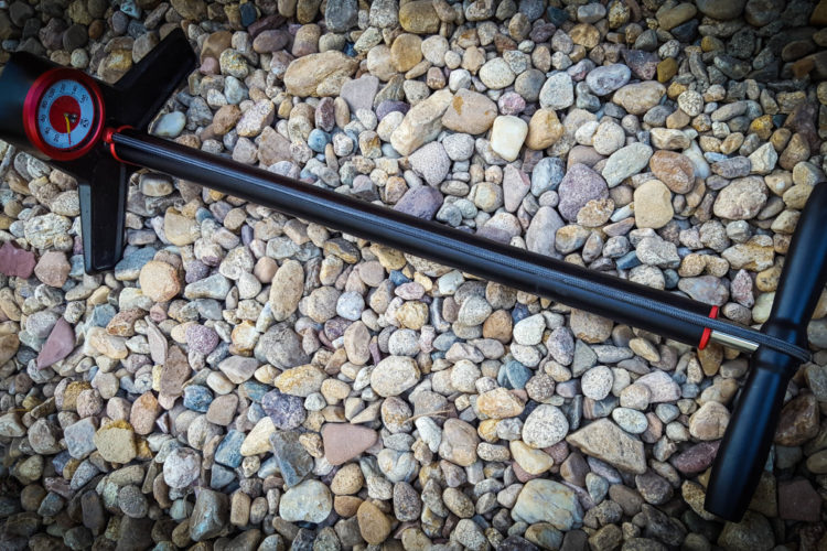 A black and red tire pressure gauge lying on a bed of small, colorful stones. The gauge features a circular dial for measuring pressure, with a long handle and a T-shaped grip.