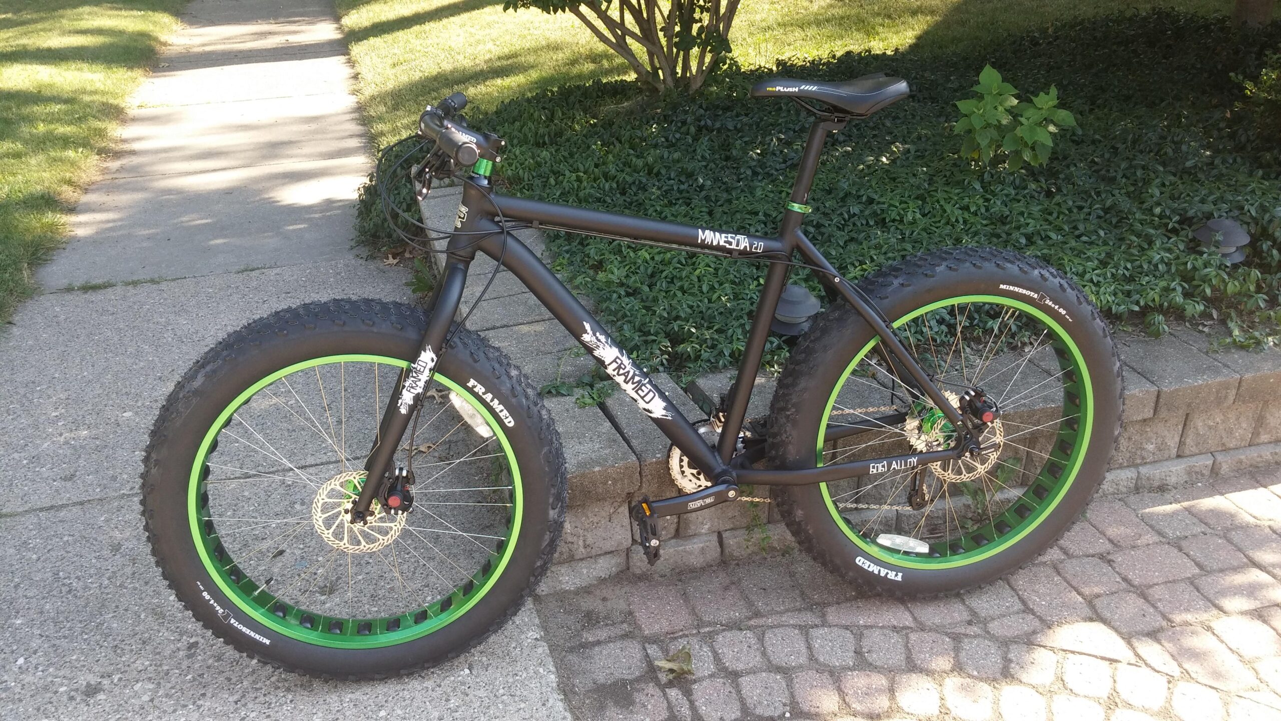 Framed Minnesota 2.0: A black fat bike with bright green accents stands on a paved pathway beside a lawn. The bike features wide tires, a sturdy frame, and disc brakes, positioned near a stone border and shrubs. Sunlight casts shadows, highlighting the bike's details.