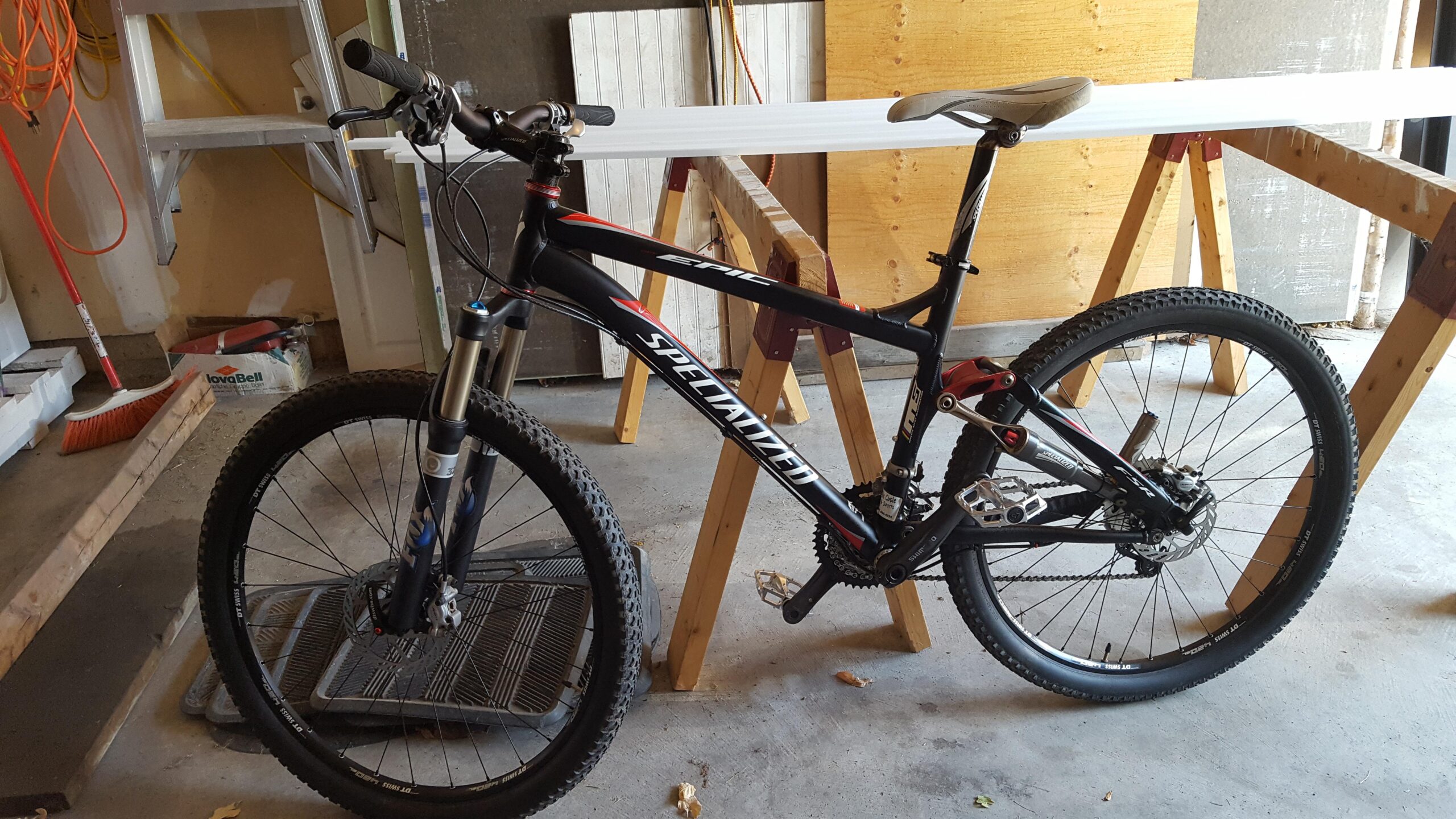 Specialized Epic FSR: A black Specialized mountain bike leaning against a wooden sawhorse in a garage. The bike features thick tires, front suspension, and a varied gear setup. The background includes a broom, some construction materials, and a partially painted wall, indicating an active workspace.