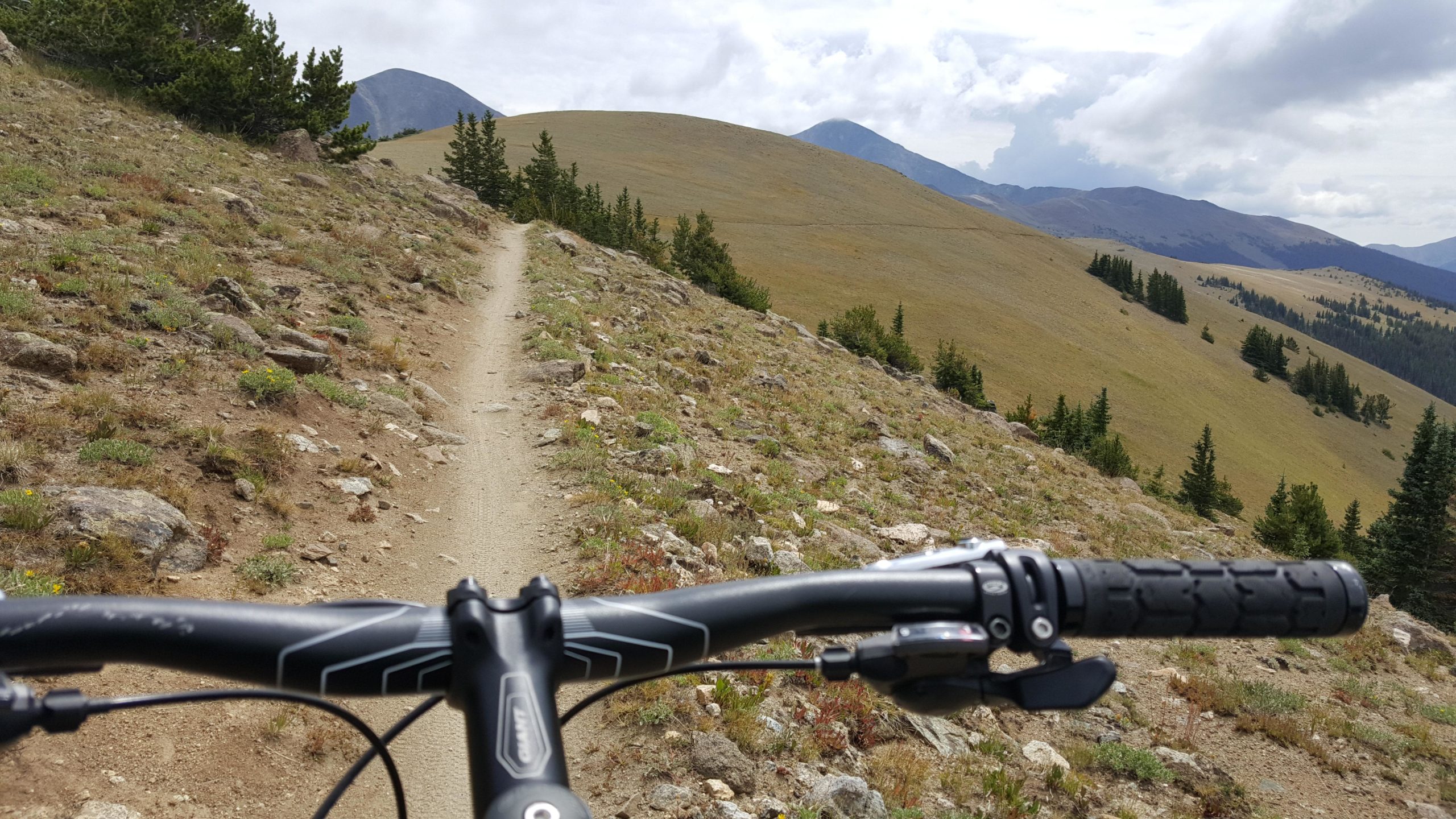 A view from the handlebars of a mountain bike on a dirt trail surrounded by rocky terrain and sparse vegetation, with rolling hills and mountains in the background under a partly cloudy sky. Monarch Crest Trail mountain bike trail.