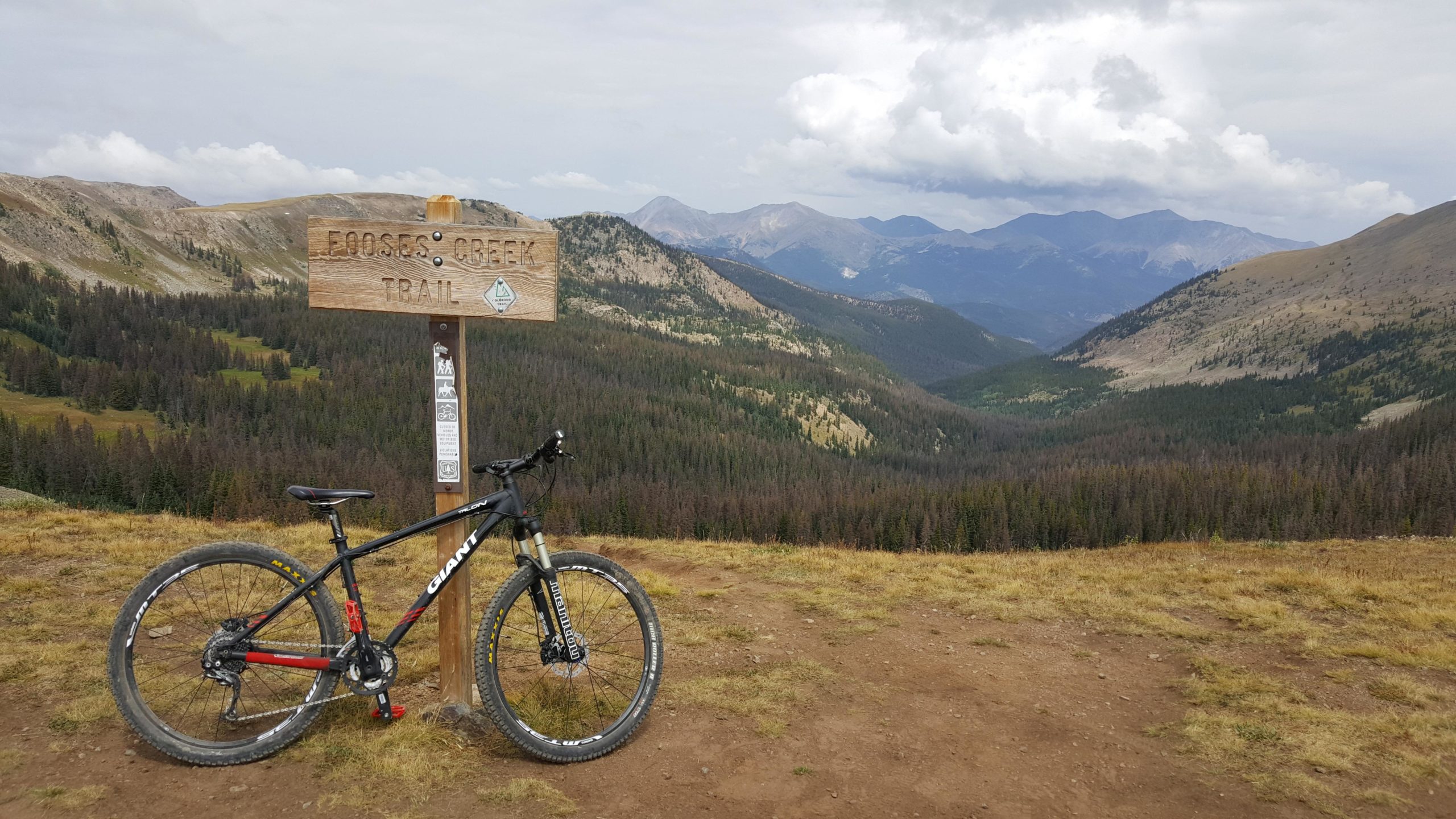 A mountain bike leaning against a wooden trail sign labeled "Mooses Creek Trail," set against a backdrop of rolling hills and cloudy skies. The landscape features a mix of green and brown vegetation, with distant mountains visible under the vast sky. Monarch Crest Trail mountain bike trail.