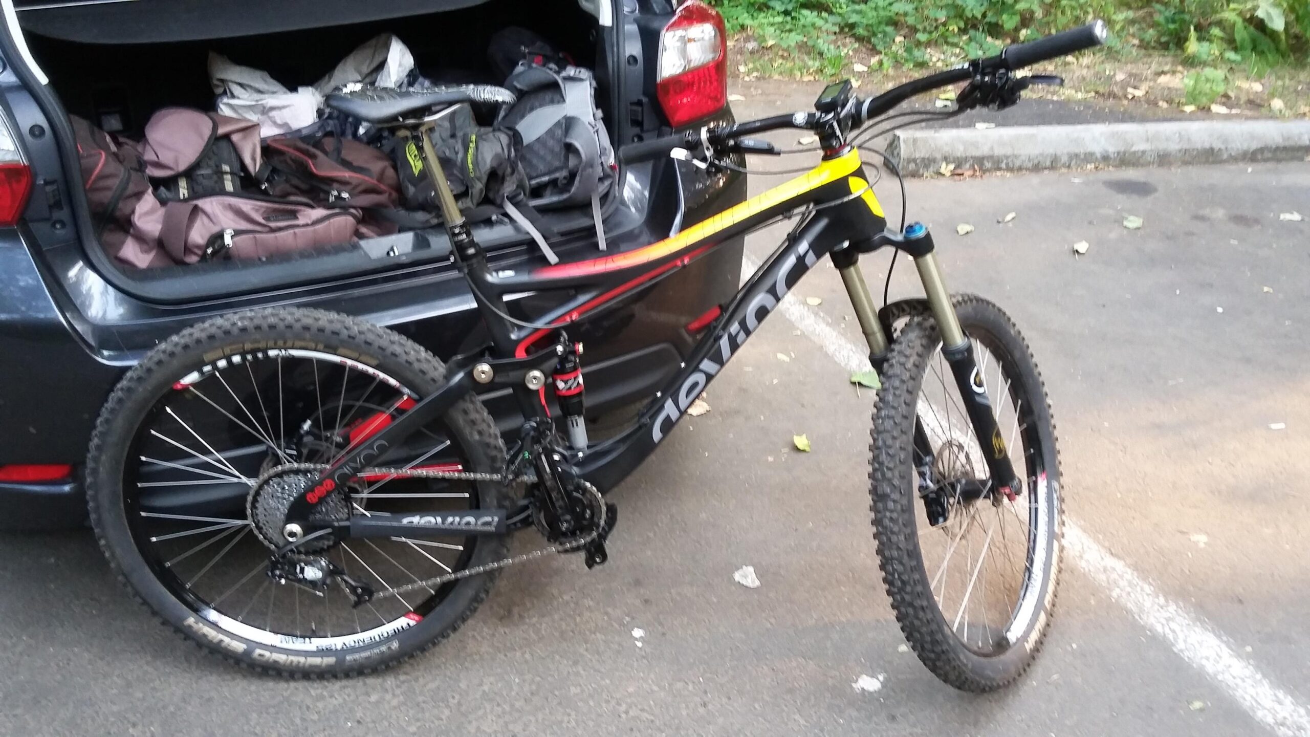 Devinci Dixon Carbon RS: A black and red mountain bike is parked near the open trunk of a car, which is filled with bags and equipment. The bike features large, knobby tires and a sturdy frame, ready for off-road adventures. The scene is set in a natural environment with greenery in the background.